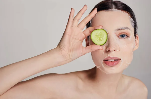 Close up girl in a moisturizing mask with a fresh cucumber on the face in the background serious with open eye and right hand holds a cucumber near face