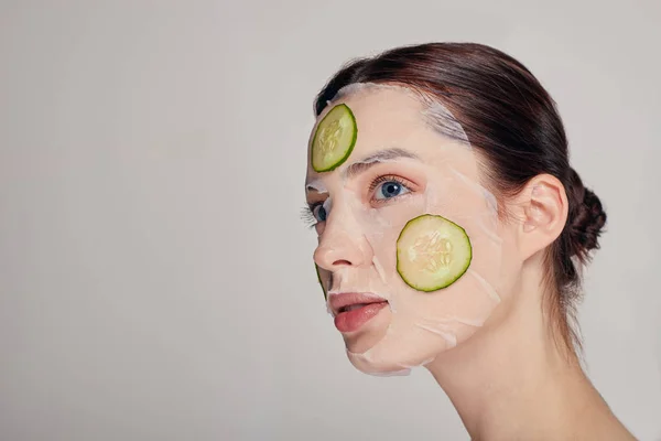 Close-up zachte geavanceerde meisje in een hydraterende masker met een frisse komkommer op het gezicht in de achtergrond kijkt weg — Stockfoto