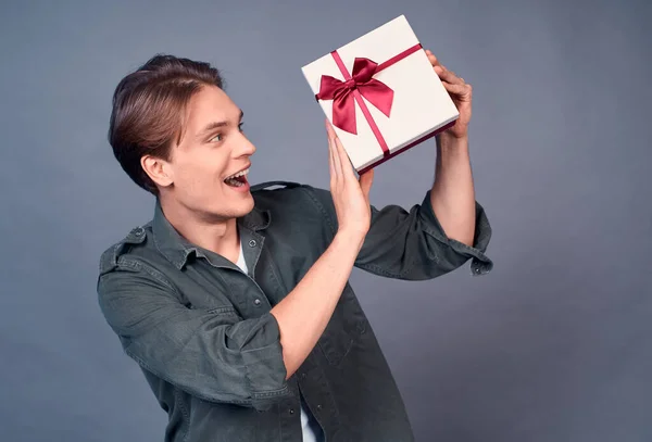 一个快乐而有趣的年轻人收到生日礼物 拿着一个包装好的盒子摇晃着身子 试着猜里面是什么 尖叫着就像打开的礼物一样兴奋而兴奋 灰色的背景 — 图库照片