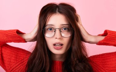 Kırmızı süveterli, pembe arka planda gözlüklü, uzun saçlı, heyecanlı esmer kadın. Sorunları ve zor durumları yakından dinlemek istemediği için ellerini başının üstüne koyuyor. Boşluğu kopyala.