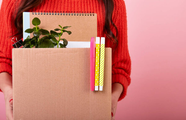 Slender fired girl in a red knitted sweater, holding a cardboard box in her hands on a pink background. In search of a new job after being fired due to quarantine. New life. Copy space.