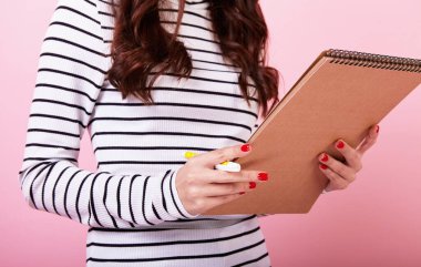 Çizgili bluzlu kırmızı manikürlü, kalemli ve defterli zarif bir esmer öğrenci. Bir bayan pembe arka planda yeni bilgiler edinmek için notlar alır. Öğrenme konsepti. Boşluğu kopyala.