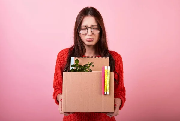 Crying dismissed young cute student in a knitted sweater and glasses. The lady gathered work items in a cardboard box to deliver them home on a pink background. New life. Copy space.