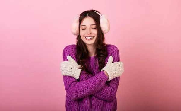 ニットのセーターとミトンの優雅な若い女性と彼女の頭の上にふわふわの冬のピンクのヘッドフォン 少女は優しく笑顔で寒さから身を抱きしめる ピンクの背景の写真 — ストック写真
