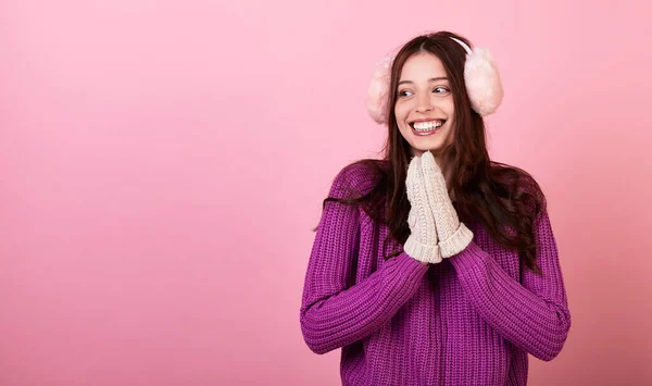 ニットのセーターとミトンの優雅な若い女性と彼女の頭の上にふわふわの冬のピンクのヘッドフォン 少女は優しく笑顔で世界平和を祈ります ピンクの背景の写真 スペースのコピー — ストック写真