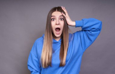 Şirin Avrupalı kadın model, uzun sarı saçlı ve kahverengi gözlü, mavi kapüşonlu, elini kafasında tutuyor ve şaşkınlıkla ağzını açıyor, şok geçiriyor..
