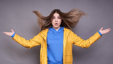 Genç büyüleyici beyaz model, mavi kazak ve sarı yağmurluk içinde, şaşırttı farklı yönlerde kollarını, onun uzun sarı saç dalgalanan rüzgar.