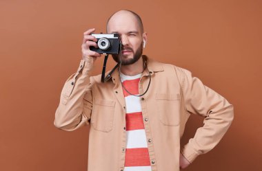 Çekici, beyaz, kel sakallı, modaya uygun çizgili tişörtlü, bej gömlekli adam, kahverengi arka planda poz veren eski bir kamerada fotoğraf çekiyor..