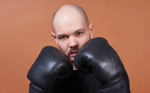 Close-up, attractive charismatic young man with beard and brown eyes, in a black T-shirt and boxing gloves, stands in a boxer rack, holds his hands near his face and confidently looks straight.