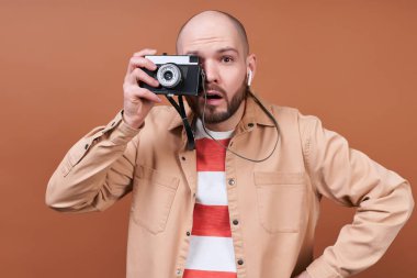 Gördüklerine hayran olan genç, kel, şık çizgili tişörtlü ve bej gömlekli bir adam kahverengi bir arka planda poz veren bir kamerayla fotoğraf çekiyor..
