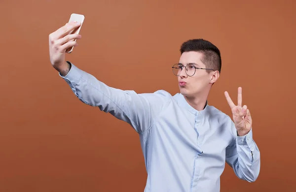 一个可爱 快乐的男人 戴着眼镜 穿着蓝色的衬衫 手里拿着一部手机 拿着一个自拍 照片上的褐色背景 — 图库照片