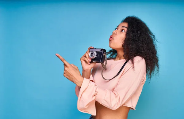 Astonished young African American girl in a nude sweater holding a retro vintage camera, pointing her index finger to the side, face surprised. Copy space.