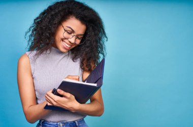 Mutlu, genç Afrikalı bir kadın, sıradan giyinmiş, mavi arka planda izole edilmiş, not defterine not alıyor. Eğitim konsepti, kız öğrenci, üniversite, üniversite. Boşluğun kopyası.