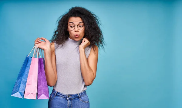 Attractive young dark skinned woman with a bunch of shopping bags worried she couldn\'t stop buying. Studio shot isolated over blue background.
