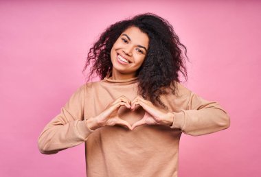Çekici, çekici, güzel, koyu tenli, siyah kıvırcık saçlı, günlük bej ceket giyen, gülümseyen ve pembe arka planda poz veren genç bir kız..