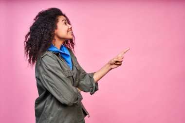 Profil tarafında koyu tenli, şaşırmış çılgın bir kızın fotoğrafı. İnanılmaz bir haberden etkilenmiş ve bir tarafı işaret ediyor. İzole edilmiş pembe kıyafetler giyiyor. Uzayı sağa kopyala.