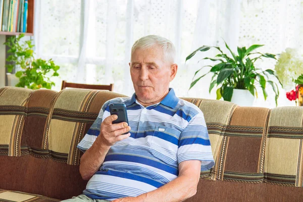 Close-up of an elderly senior man with gray hair sitting on a sofa and holding a mobile phone. The concept of using modern technologies for the elderly.