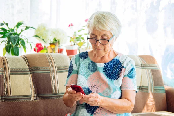 Close-up of an elderly woman with glasses and gray hair sitting on a sofa and holding a mobile phone. The concept of using modern technologies for the elderly.