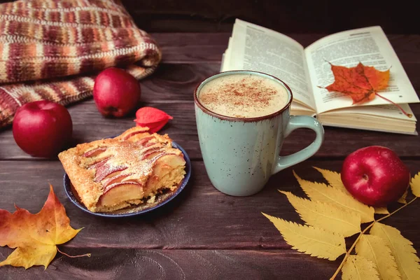 Apple pie, a book, yellow leaves, red apples, and a Cup of coffee on a dark wooden table. Autumn background, time for tea and reading.
