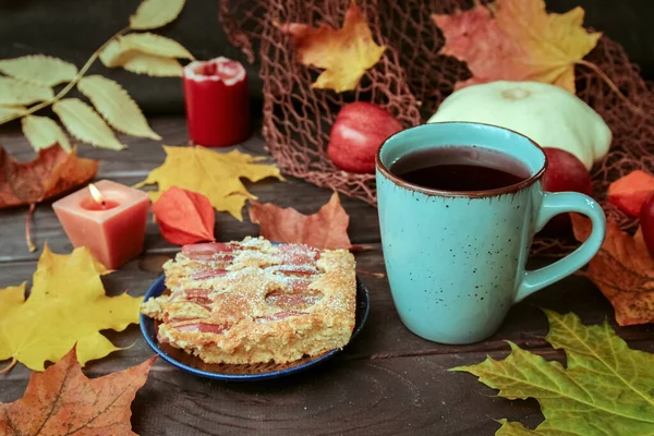 A piece of cake and a Cup of tea on a dark wooden table with autumn leaves, red apples and burning candles. Autumn background, time for tea. The concept of peace and harmony.