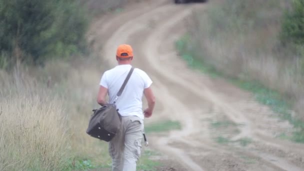 村の道でヒッチハイクをしている男 若いあごひげの男が野原で車を止める 旅行仲間 旅行者 放浪者 — ストック動画