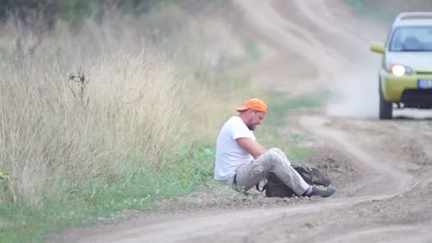 一个男人在乡村公路上搭便车 一个留着胡子的年轻男子在田野里拦住一辆汽车 旅行伴侣 旅行者 流浪者 — 图库视频影像