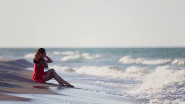 若い女の子が海岸に一人で座っている 赤いドレスの若い女性がビーチに座っている — ストック動画