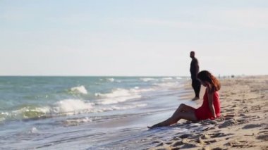 Genç bir adam deniz kıyısında oturan kırmızı elbiseli bir kızın yanından geçiyor.. 