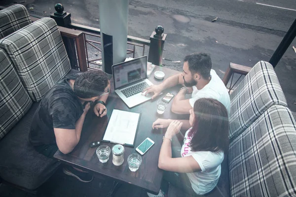 Side view and top. The company of three businessmen are discussing at the table in coffee, writing in a notebook, looking at a laptop, satkan with water, car keys, magazines, a smartphone, online transactions.