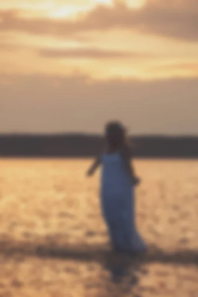 Homályos - szőke lány séta a vízen a naplemente fehér ruhát. — Stock Fotó
