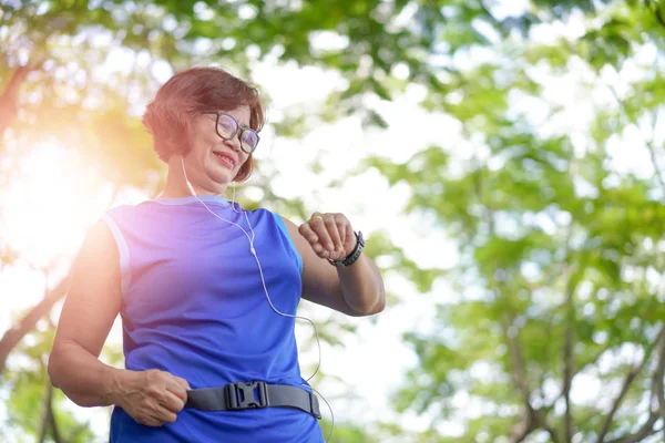 上級アジアの女性はスマートな時計から時間をチェックします 公園でジョギング中の心拍数をチェックします スマートウォッチを走りながら彼女のハートビート — ストック写真