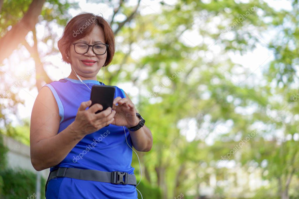 Happy senior asian woman holding smart phone with listening to music. Checking heart rate while jogging in the park. Looking on mobile phone her heartbeat while running   