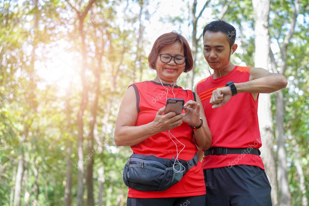Happy senior asian woman with man or personal trainer checking time from smart watch. Checking heart rate while jogging in the park. Looking on smartwatch her heartbeat while running, Elderly care
