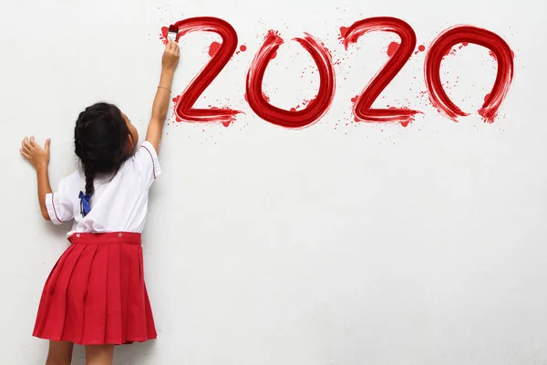 Little girl holding a paint brush painting happy new year 2020 on a white wall — Stock Photo, Image