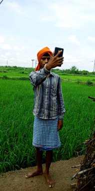 DISTRICT KATNI, INDIA - 14 Ağustos 2019: Bir Hintli köy çiftçisi tarım yeşil çim tarlası çevresindeki akıllı telefondan video çağrısı yapıyor.