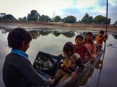 DISTRICT KATNI, INDIA - HAZİRAN 01, 2020: Nehirde bir grup çocukla birlikte yelken açarken dizüstü bilgisayar kullanan Hintli bir balıkçı çocuk, köylüler için dijital öğrenme.