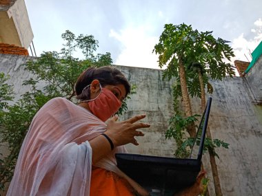 DISTRICT KATNI, INDIA - 16 Mayıs 2020: Hintli bir ofis kızı evde dizüstü bilgisayar teknolojisi kullanıyor ve Corona virüsü için yüz maskesi takıyor.