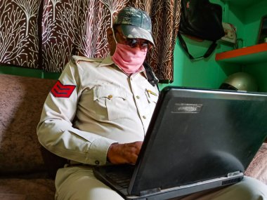 DISTRICT KATNI, INDIA - 21 Mayıs 2020: Evde dizüstü bilgisayarda çalışan Hintli bir polis Coronavirus koruması için maske takıyor.