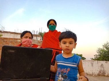 DİSTRİK KATNI, INDIA - 23 Mayıs 2020: Hintli küçük bir çocuk dizüstü bilgisayar tutuyor ve annesi açık arka planda otururken kameraya bakıyor.