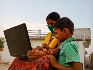 DISTRICT KATNI, INDIA - 23 Mayıs 2020: Oğluyla birlikte açık alanda dizüstü bilgisayar teknolojisi öğrenen Hintli fakir bir kadın Corona virüsü koruması için yüz maskesi takıyor.