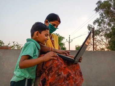 DISTRICT KATNI, INDIA - 23 Mayıs 2020: Oğluyla birlikte açık alanda dizüstü bilgisayar teknolojisi öğrenen Hintli fakir bir kadın Corona virüsü koruması için yüz maskesi takıyor.