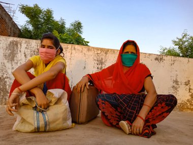 DISTRICT KATNI, INDIA - 21 Mayıs 2020: Hindistan köyünün anne ve kızı Corona virüsü koruması için yüz maskesi takıyor ve arka planda bavullarıyla birlikte oturuyorlar.