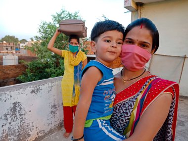 DISTRICT KATNI, INDIA - 21 Mayıs 2020: Corona virüsü koruması için yüz maskesi takan Hintli bir anne oğlunu arka planda kucağına aldı.