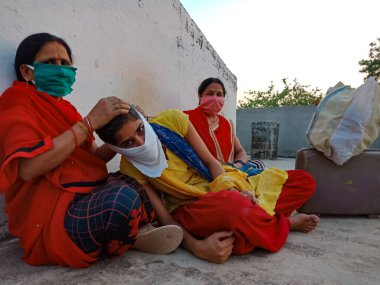 DISTRICT KATNI, INDIA - 21 Mayıs 2020: Korona virüsü koruması için yüz maskesi takan Asyalı köylü kadınlar gökyüzü duvarı arka planında bavullarıyla birlikte oturuyorlar.