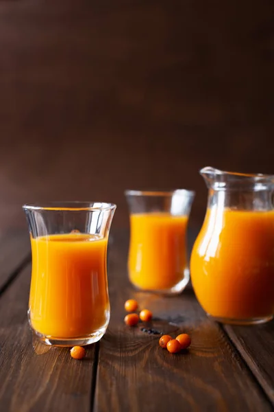Sea buckthorn drink in glasses and jug on a wooden background. Scattered around berry