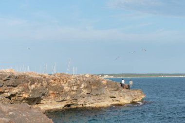 Sa Rapita, Campos, Mayorka, İspanya - 25 Mayıs 2020: Arka planda iki balıkçının uçan martılara baktığı bir koyda kıyı manzarası