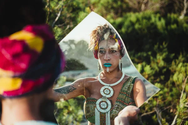 Woman in wild nature, with African face paint and ethnic clothes looking at the mirror