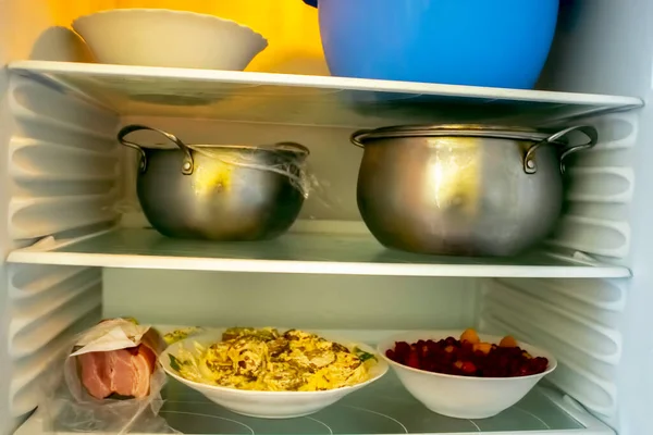 Shelves of an open frige refrigerator with different food, cooked food in pot and plastic container.