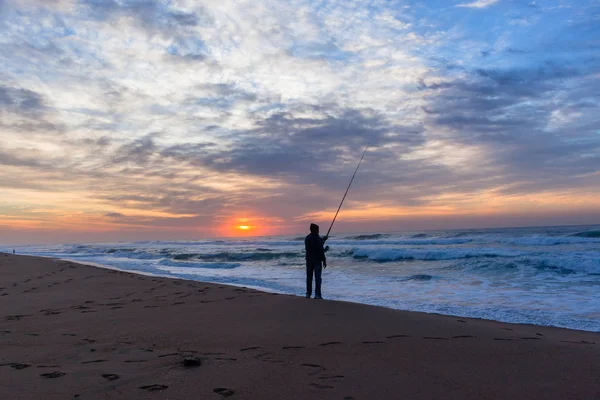 Fisherman fishing silhouetted unidentified on beach waters edge with ocean waves early morning dawn sunrise.