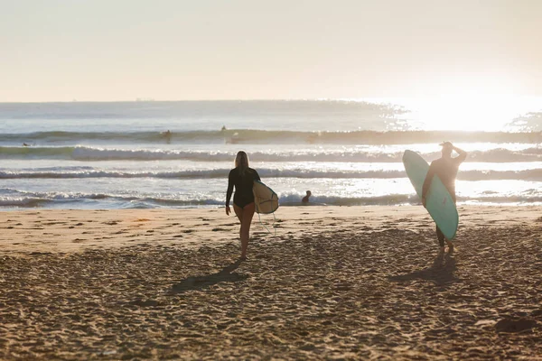 海の朝の日光のまぶしさに向かってビーチを歩いて女の子男の子朝をボード長い間サーフィンに行く — ストック写真